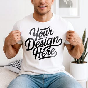 May include: White t-shirt with the text "Your Design Here" in a black script font. The t-shirt is being held up by a person. The person is wearing blue jeans.