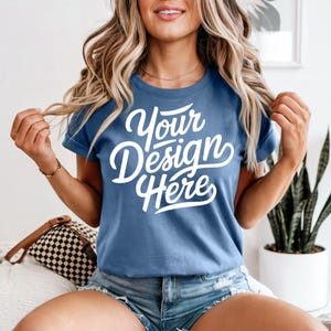 May include: A blue t-shirt with the text "Your Design Here" in white script. The shirt has short sleeves and a crew neck. The model is wearing denim shorts and a gold necklace.