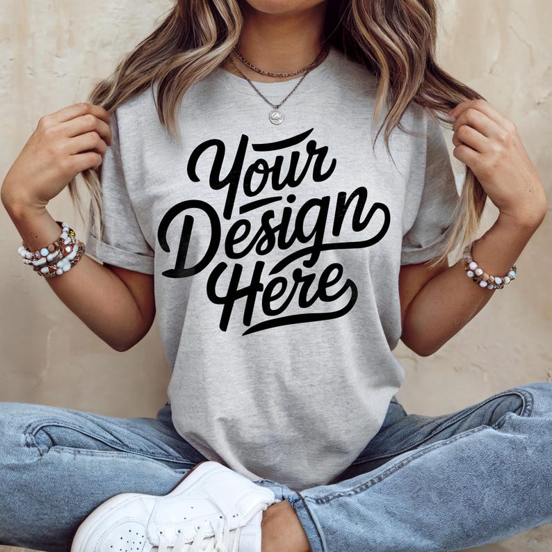 May include: A light gray t-shirt with the text "Your Design Here" in a black script font. The shirt has short sleeves and is being worn with blue jeans and white sneakers. The model is wearing bracelets and a necklace.