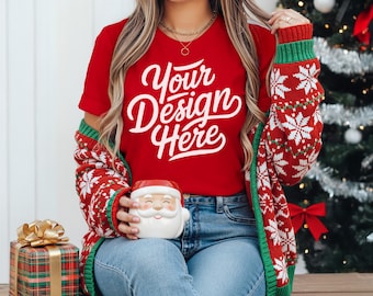 May include: A red V-neck t-shirt with the text "Your Design Here" is paired with a red, white, and green snowflake patterned cardigan. A Santa Claus mug and a plaid gift box with a gold bow are also visible. The theme is Christmas.