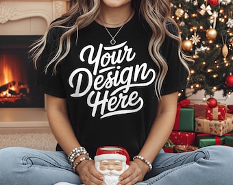 May include: Black t-shirt with the text "Your Design Here" in white script. The person is holding a Santa Claus figurine. The background includes a Christmas tree and a fireplace, suggesting a holiday theme.