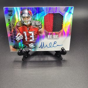 May include: A silver and holographic football card featuring Mike Evans, a wide receiver for the Tampa Bay Buccaneers. The card has a patch of Evans' jersey, a rookie card designation, and his autograph. The card is on a black stand.
