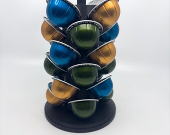 Op de afbeelding: Een zwarte draaibaar standaard houdt een verscheidenheid aan kleurrijke koffie capsules vast. De capsules zijn in een cirkelvormige opstelling geplaatst en zijn een mix van blauw, groen en goud.
