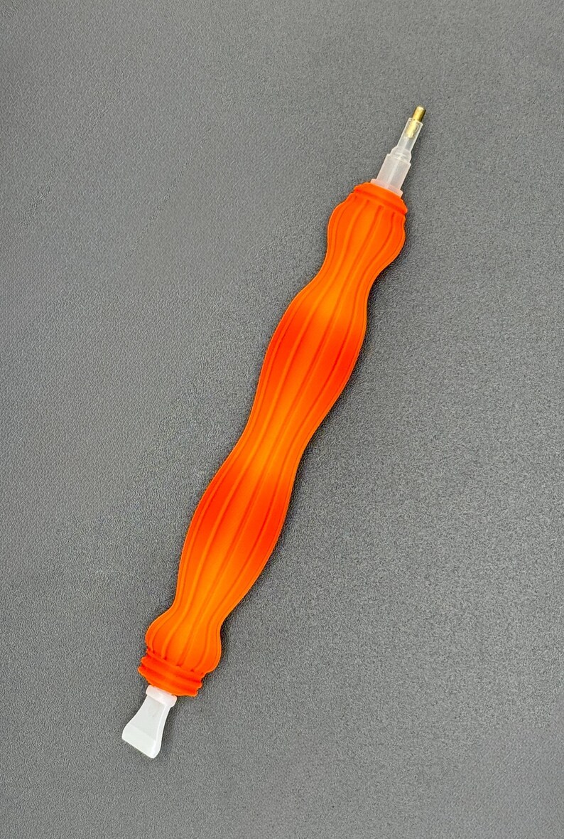 May include: An orange and yellow diamond painting pen with a ribbed handle. The pen has a clear plastic tip and a gold-coloured metal tip. The pen is designed for precision and detail work in diamond painting projects.