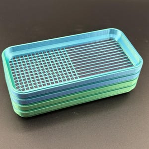 May include: A rectangular, multi-layered tray in shades of green and blue. The top layer features a grid pattern on one half and a striped pattern on the other. The tray is likely made of plastic or a similar material.
