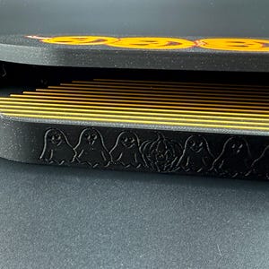 May include: A black Halloween-themed box with a lid featuring three orange jack-o'-lantern faces. The box's side is decorated with ghost and pumpkin silhouettes. Inside, there are multiple yellow and black striped dividers.