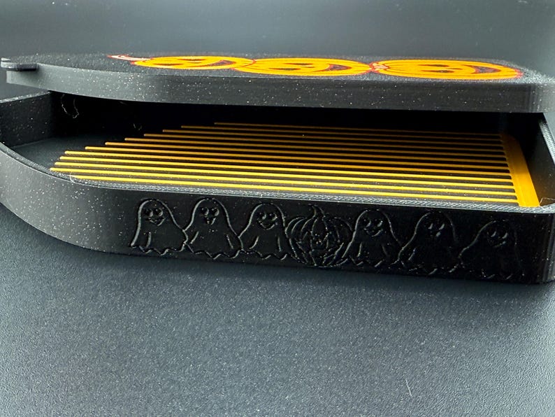May include: A black Halloween-themed box with a lid featuring three orange jack-o'-lantern faces. The box's side is decorated with ghost and pumpkin silhouettes. Inside, there are multiple yellow and black striped dividers.