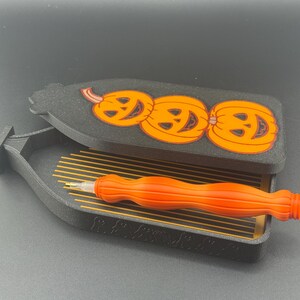 May include: A black and orange Halloween-themed storage box with a lid featuring three smiling jack-o'-lanterns. Inside the box is an orange and white craft tool, designed for diamond painting.