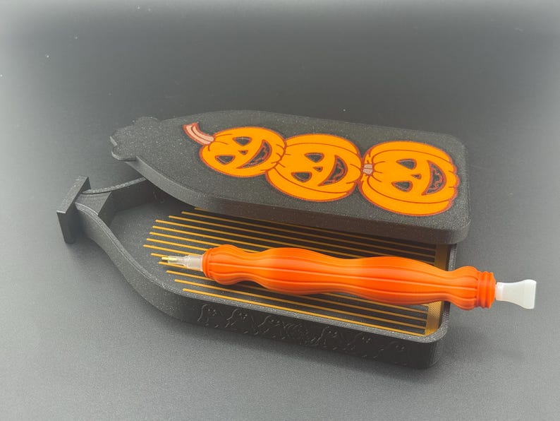 May include: A black and orange Halloween-themed storage box with a lid featuring three smiling jack-o'-lanterns. Inside the box is an orange and white craft tool, designed for diamond painting.
