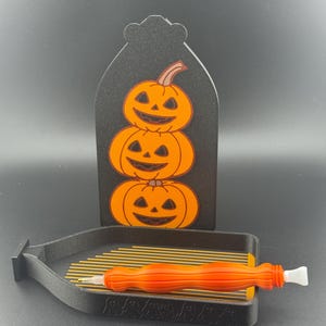 May include: A black Halloween decoration with three stacked orange pumpkins with carved faces. A black tray with gold rods and an orange stylus are in the foreground. The pumpkins are on a black tombstone-shaped background.