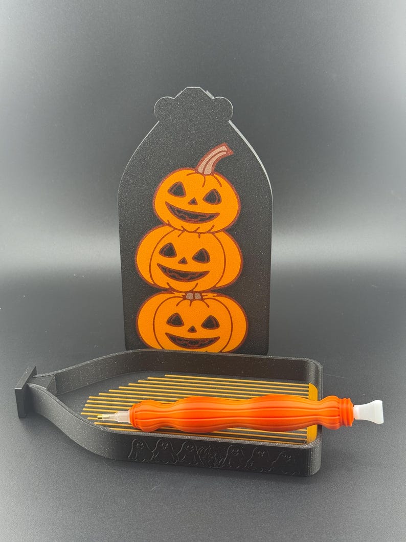 May include: A black Halloween decoration with three stacked orange pumpkins with carved faces. A black tray with gold rods and an orange stylus are in the foreground. The pumpkins are on a black tombstone-shaped background.