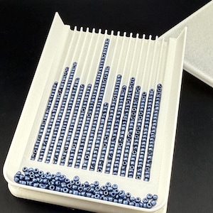 May include: A white bead sorting tray filled with rows of blue beads. The tray has a ridged design and is partially filled with loose beads. A white lid is in the background. The beads are small and cylindrical.