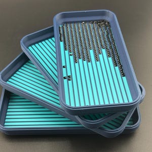 May include: Three stacked, rectangular trays with a teal and navy blue color scheme. The trays have a striped pattern on the inside and are filled with small, dark objects. The trays are likely used for organizing small items.