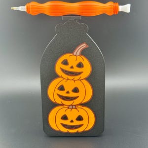 May include: A black Halloween-themed diamond painting pen holder featuring three stacked orange pumpkins. An orange pen rests on top. The pen holder is a decorative accessory for diamond painting.