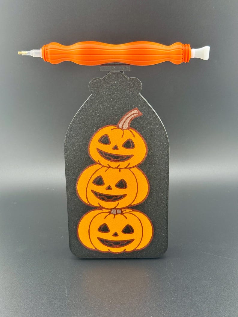 May include: A black Halloween-themed diamond painting pen holder featuring three stacked orange pumpkins. An orange pen rests on top. The pen holder is a decorative accessory for diamond painting.