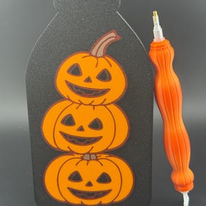 May include: A black Halloween decoration with three orange jack-o'-lanterns stacked on top of each other. An orange diamond painting pen is to the right. The pumpkins have carved faces and brown stems. The background is black.