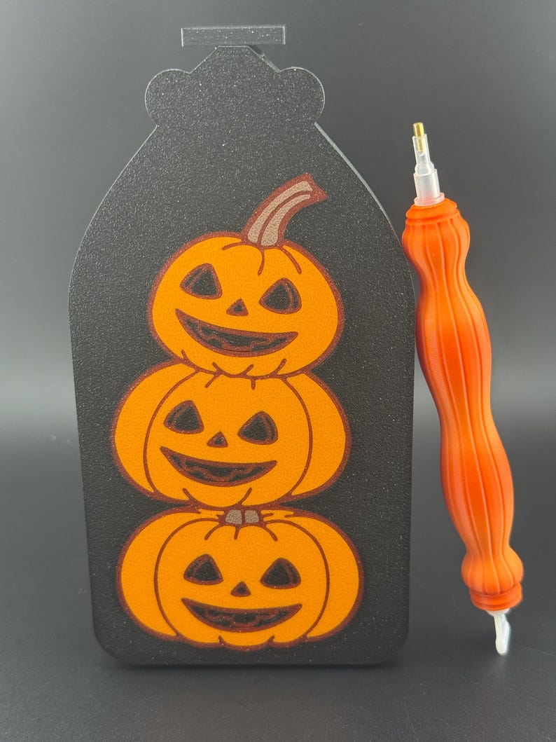 May include: A black Halloween decoration with three orange jack-o'-lanterns stacked on top of each other. An orange diamond painting pen is to the right. The pumpkins have carved faces and brown stems. The background is black.
