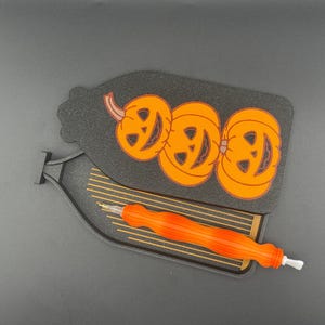 May include: Black Halloween-themed craft tool holder with three orange jack-o'-lanterns. An orange and white craft tool rests in the holder. The holder is shaped like a bottle.