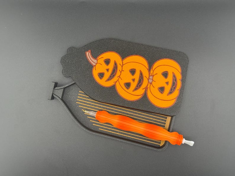 May include: Black Halloween-themed craft tool holder with three orange jack-o'-lanterns. An orange and white craft tool rests in the holder. The holder is shaped like a bottle.