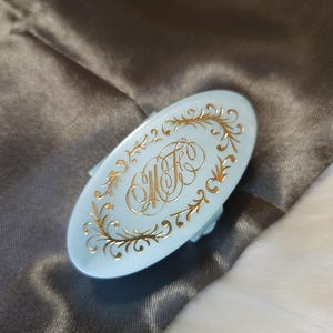 May include: A light blue oval-shaped trinket box with a gold engraved design featuring a monogram and floral accents.