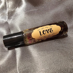 May include: A clear glass roll-on bottle with a black cap, filled with a liquid and dried petals. A tan label with the word "Love" in black script is affixed to the bottle. The bottle is resting on a grey, textured surface.