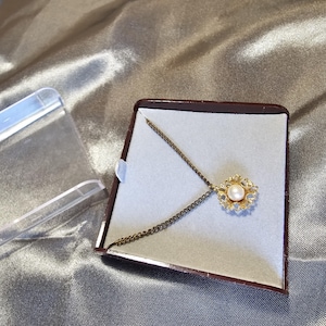 Puede incluir: Un collar de oro con un colgante de flor de perla y strass en una caja de terciopelo marrón.