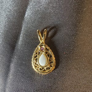 May include: A gold-colored pendant with a teardrop shape. The pendant features a white stone in the center, surrounded by an ornate filigree design. A small pink stone is set at the top.