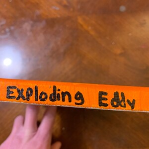K&ouml;nnte beinhalten: Nahaufnahme einer orangefarbenen Leinwand mit den Worten "Exploding Eddy" in schwarzer Farbe.