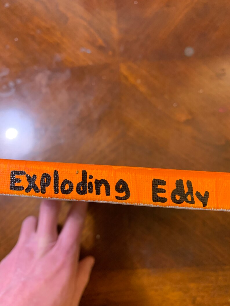 K&ouml;nnte beinhalten: Nahaufnahme einer orangefarbenen Leinwand mit den Worten "Exploding Eddy" in schwarzer Farbe.