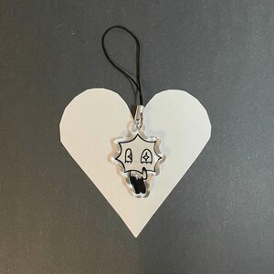 May include: A clear acrylic charm with a black and white cartoon skull design, hanging from a black cord. The charm is attached to a white heart-shaped paper cutout, set against a dark gray background.