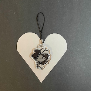May include: A clear acrylic charm featuring a cartoon character with dark hair and a sword. The charm is attached to a black cord and is placed on a white heart-shaped cutout against a gray background.