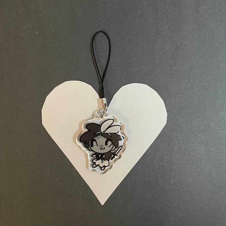 May include: A clear acrylic charm featuring a cartoon character with dark hair and a sword. The charm is attached to a black cord and is placed on a white heart-shaped cutout against a gray background.