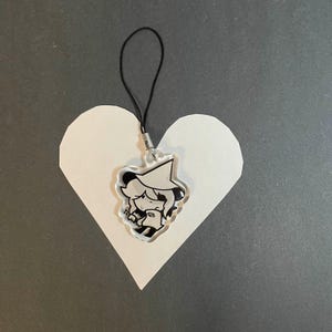 May include: A clear acrylic charm featuring a black and white illustration of a character wearing a witch hat. The charm hangs from a black cord and is set against a white heart shape on a gray background.