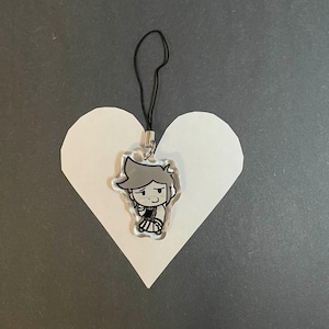May include: A clear acrylic charm featuring a cartoon character with gray hair and a black and white outfit. The charm is attached to a black lanyard. The charm is placed on a white heart shape against a gray background.