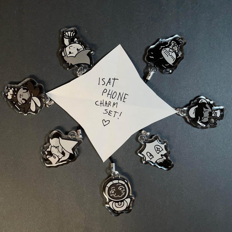 May include: A set of seven black and white phone charms with various cartoon character designs. A white note in the center reads "ISAT PHONE CHARM SET!" with a heart symbol. The charms are arranged around the note.