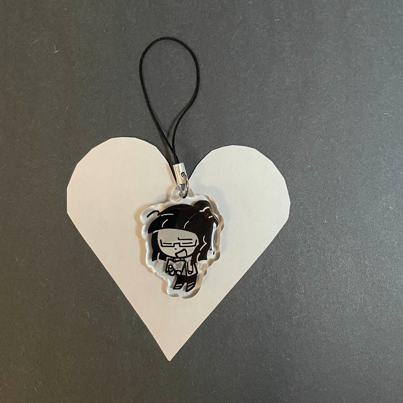 May include: A clear acrylic charm featuring a black and white cartoon character with glasses and dark hair, attached to a black cord. The charm is placed on a white heart-shaped cutout against a gray background.