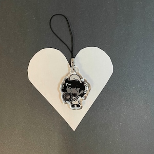 May include: A clear acrylic charm featuring a cartoon character with black hair and a chef's hat, holding a pan. The charm hangs from a black cord. The charm is placed on a white heart-shaped cutout against a gray background.