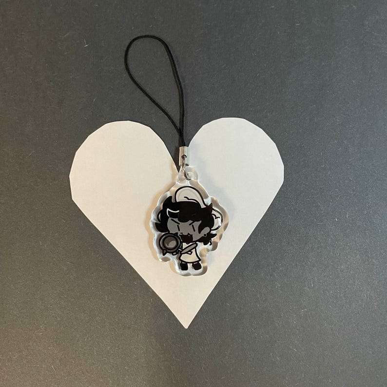 May include: A clear acrylic charm featuring a cartoon character with black hair and a chef's hat, holding a pan. The charm hangs from a black cord. The charm is placed on a white heart-shaped cutout against a gray background.