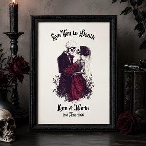 May include: Black framed art print depicting a skeletal couple embracing, with the text "Love You to Death." The couple is surrounded by red roses, with the names "Liam & Maria" and the date "21st June 2026" below. The frame is black.
