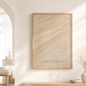 Puede incluir: Un cuadro rectangular con marco de madera clara que presenta un patr&oacute;n beige de textura similar a la piedra con l&iacute;neas diagonales y motas oscuras. La palabra GROUNDED est&aacute; impresa en letras may&uacute;sculas peque&ntilde;as y oscuras en la parte inferior.