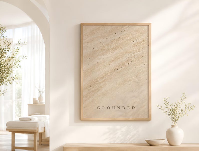 Puede incluir: Un cuadro rectangular con marco de madera clara que presenta un patr&oacute;n beige de textura similar a la piedra con l&iacute;neas diagonales y motas oscuras. La palabra GROUNDED est&aacute; impresa en letras may&uacute;sculas peque&ntilde;as y oscuras en la parte inferior.