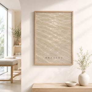 Puede incluir: Una impresi&oacute;n de arte mural rectangular en un marco de madera clara con una fotograf&iacute;a de agua brillante bajo el sol en tonos beige y crema. La palabra BREATHE est&aacute; impresa en una fuente sans-serif oscura y sencilla en la parte inferior.