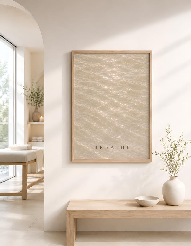 Puede incluir: Una impresi&oacute;n de arte mural rectangular en un marco de madera clara con una fotograf&iacute;a de agua brillante bajo el sol en tonos beige y crema. La palabra BREATHE est&aacute; impresa en una fuente sans-serif oscura y sencilla en la parte inferior.