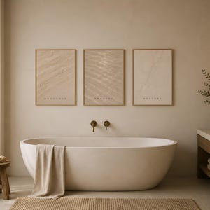 Puede incluir: Tres l&aacute;minas de pared rectangulares en marcos de madera clara colgadas sobre una ba&ntilde;era ovalada blanca. Las l&aacute;minas presentan texturas abstractas en beige y las palabras GROUNDED, BREATHE y RESTORE en letras may&uacute;sculas oscuras.