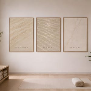 Puede incluir: Tres l&aacute;minas rectangulares de pared en marcos de madera clara, cada una con una textura abstracta en tonos beige. La l&aacute;mina de la izquierda muestra la palabra GROUNDED, la central BREATHS y la de la derecha la palabra RESTORE.