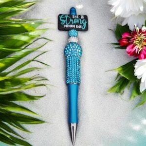 May include: A blue pen with a turquoise rhinestone tassel and a black sign that reads "She is Strong Proverbs 31:25" on top.