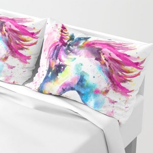 May include: Two white pillowcases with a watercolor painting of a unicorn's head. The unicorn has a pink mane and a blue horn. The background is white with splashes of pink, blue, and yellow paint.