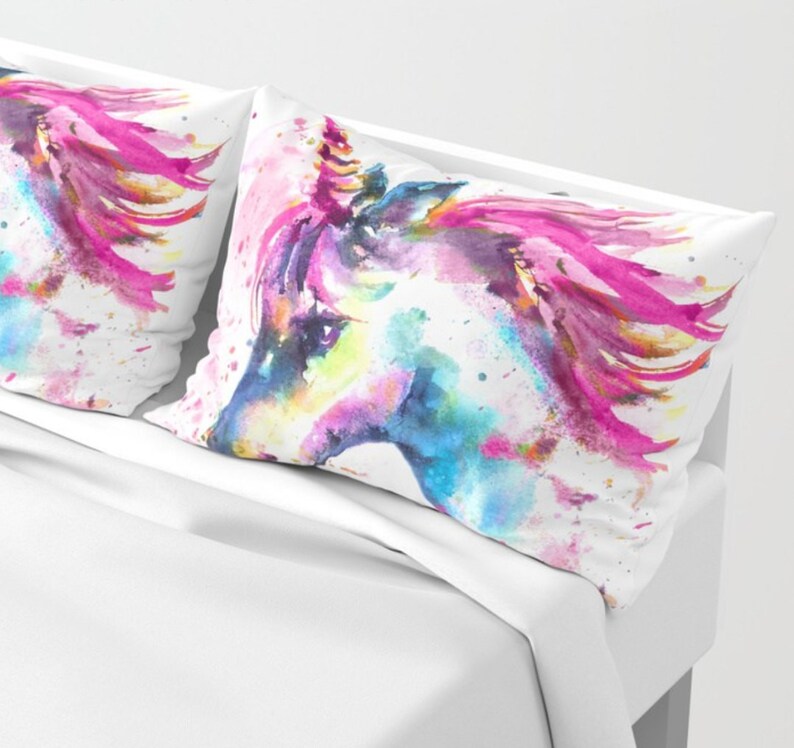 May include: Two white pillowcases with a watercolor painting of a unicorn's head. The unicorn has a pink mane and a blue horn. The background is white with splashes of pink, blue, and yellow paint.
