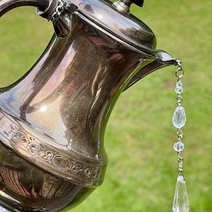 Puede incluir: Una tetera de metal plateado con una banda ornamental en relieve alrededor del cuerpo, un remate negro en la tapa y una cadena decorativa colgante de cuentas de vidrio facetado transparente y un colgante de cristal puntiagudo en el pico.