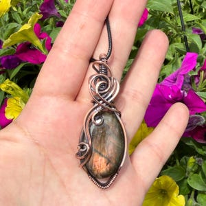 May include: A wire-wrapped pendant featuring an oval-shaped labradorite gemstone with iridescent flashes of orange and brown. The stone is encased in polished copper wire, intricately coiled and looped at the top, suspended from a black cord.
